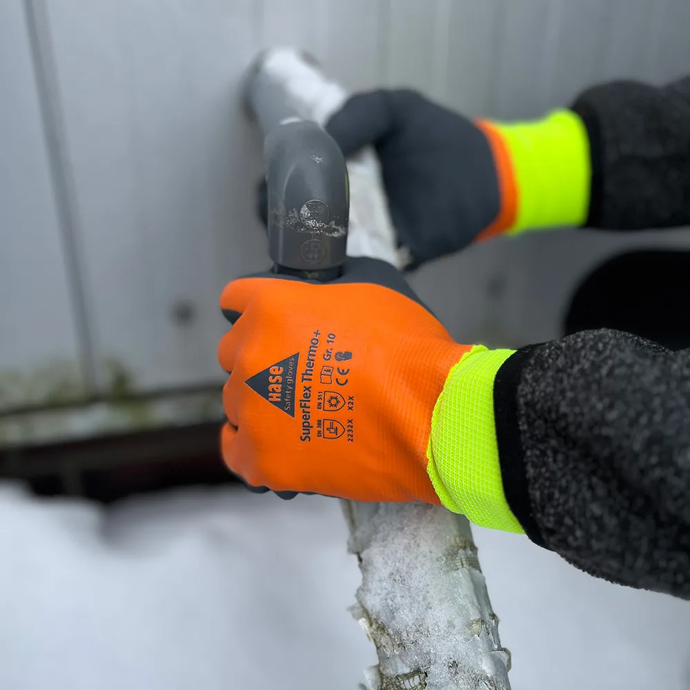 Orange HASE Kältehandschuhe SuperFlex Thermo+ greifen ein vereistes Rohr an einer Außeneinheit im Schnee