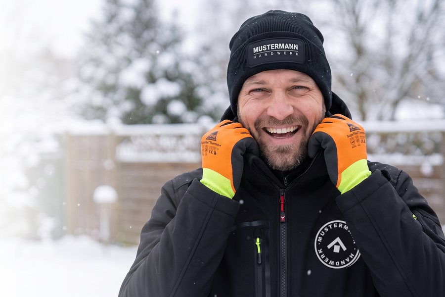 Lachender Mann mit Mütze mit Firmenlogo und orangen Winterhandschuhen in der Winterjacke, Bild KI-generiert
